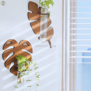 Puede incluir: Dos jardineras de pared de madera con forma de hoja con jarrones de tubos de ensayo de vidrio. Las hojas de madera marrón están montadas en una pared blanca, cada una con una planta con hojas verdes. El texto "AAHomeCrafts" es visible.