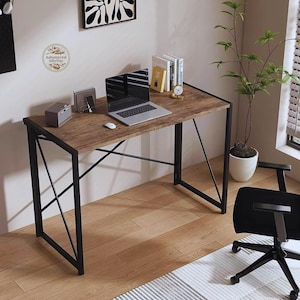 May include: A desk with a wooden top and a black metal frame. A laptop, books, and a small clock are on the desk. A plant in a white pot and a black office chair are nearby. The desk has a rustic wood grain finish.