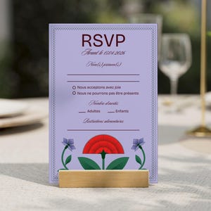 以下が含まれることがあります： ラベンダー色のRSVPカード。バーガンディ色の「RSVP」の文字と花のアクセントが特徴。名前、出欠、人数などを記入する欄があり、木製のスタンドで支えられています。テーブルの上に置かれたカード。