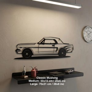 以下が含まれることがあります： クラシックマスタングのコンバーチブルを描いた黒い金属製の壁掛けアート。明るい色の壁に取り付けられています。「Classic Mustang」の文字が下にあり、サイズはMedium - 50x15 cm、Large - 70x21 cmです。時計とアクセサリー付きの棚も表示されています。