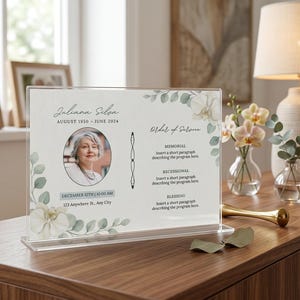 Könnte beinhalten: Ein klarer Gedenk-Acrylschild mit dem Namen "Juliana Silva" und den Daten August 1950 - Juni 2024. Das Schild enthält ein Foto, Veranstaltungsdetails und florale Akzente. Das Schild wird auf einer Holzoberfläche angezeigt.