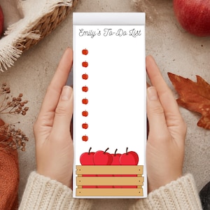 May include: A white rectangular notepad with the text Emily's To-Do List at the top, featuring a vertical column of small red apple icons for bullet points and a graphic of red apples in a wooden crate at the bottom of the page.