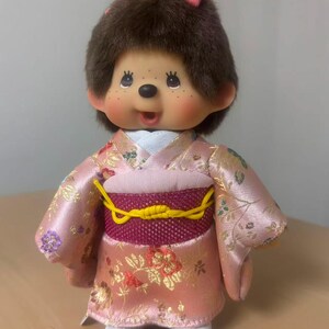 Puede incluir: Muñeca de peluche con pelo castaño, vestida con un kimono rosa floral con un obi amarillo. La muñeca tiene una cara sonriente, mejillas rosadas y un pequeño lazo rosa en la cabeza. Zapatos blancos y cola marrón.