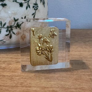 May include: A clear, rectangular block encases a gold-colored tile with floral and character designs. The tile features the number "1" and a Chinese character. The block sits on a wooden surface.