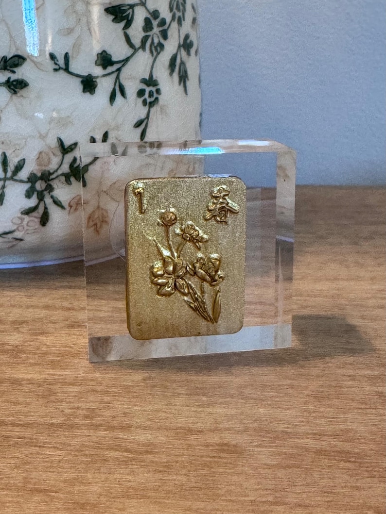 May include: A clear, rectangular block encases a gold-colored tile with floral and character designs. The tile features the number "1" and a Chinese character. The block sits on a wooden surface.
