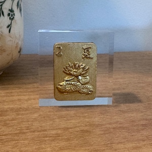 May include: A gold-colored card with the number "2" and Chinese characters, featuring a lotus flower design. The card is displayed in a clear acrylic block, showcasing its intricate details and craftsmanship. The card is likely part of a game or decorative item.
