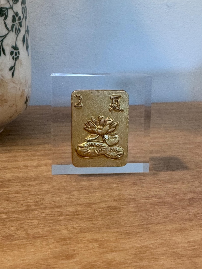 May include: A gold-colored card with the number "2" and Chinese characters, featuring a lotus flower design. The card is displayed in a clear acrylic block, showcasing its intricate details and craftsmanship. The card is likely part of a game or decorative item.