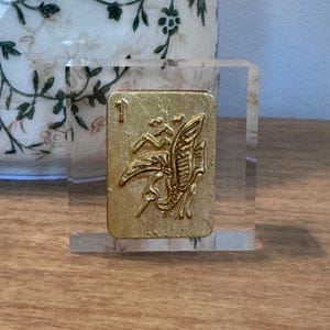 May include: A gold-colored, rectangular tile with an embossed design of a bird and foliage, set in a clear acrylic block. The tile is marked with the number 1. The background includes a decorative vase with a floral pattern.