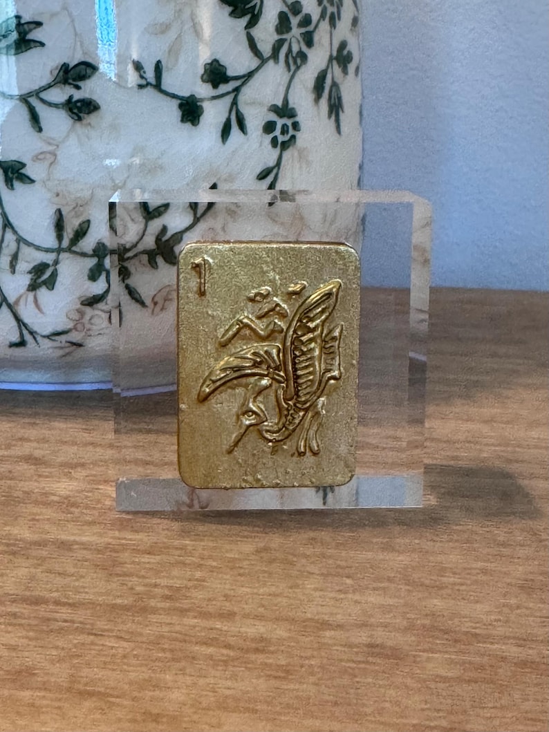 May include: A gold-colored, rectangular tile with an embossed design of a bird and foliage, set in a clear acrylic block. The tile is marked with the number 1. The background includes a decorative vase with a floral pattern.