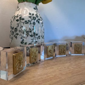 May include: Five clear acrylic blocks with gold-colored rectangular inserts featuring embossed floral designs. The blocks are arranged in a row on a wooden surface. A decorative vase with green floral patterns and yellow tulips is in the background.
