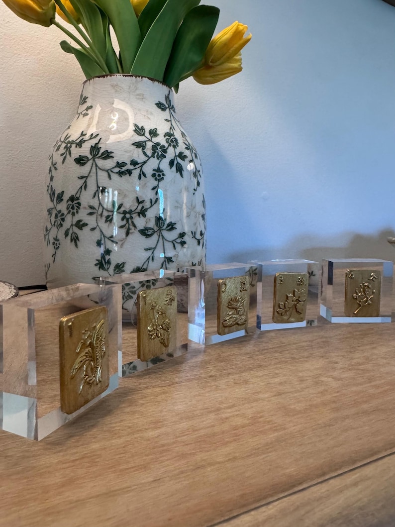 May include: Five clear acrylic blocks with gold-colored rectangular inserts featuring embossed floral designs. The blocks are arranged in a row on a wooden surface. A decorative vase with green floral patterns and yellow tulips is in the background.