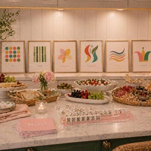 May include: A marble countertop displays a spread of food and drinks, including a charcuterie board, fruit, and olives. Six framed art prints with colorful abstract designs hang on the wall. A pink Mahjong set and napkins are on the counter.