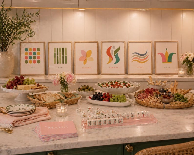 May include: A marble countertop displays a spread of food and drinks, including a charcuterie board, fruit, and olives. Six framed art prints with colorful abstract designs hang on the wall. A pink Mahjong set and napkins are on the counter.