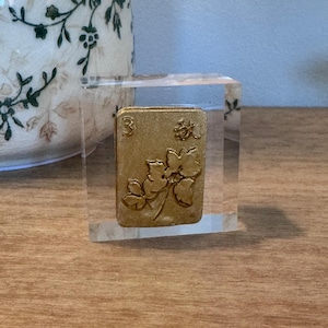May include: A gold-colored Mahjong tile with the number 3 and floral designs, encased in a clear acrylic block. The tile is set on a wooden surface, with a decorative vase in the background.