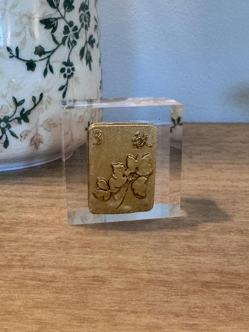 May include: A gold-colored Mahjong tile with the number 3 and floral designs, encased in a clear acrylic block. The tile is set on a wooden surface, with a decorative vase in the background.