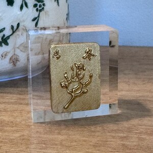 May include: A gold-colored Mahjong tile, featuring the number 4 and Chinese characters, is encased in a clear acrylic block. The tile has a floral design. The block sits on a wooden surface, with a decorative vase in the background.