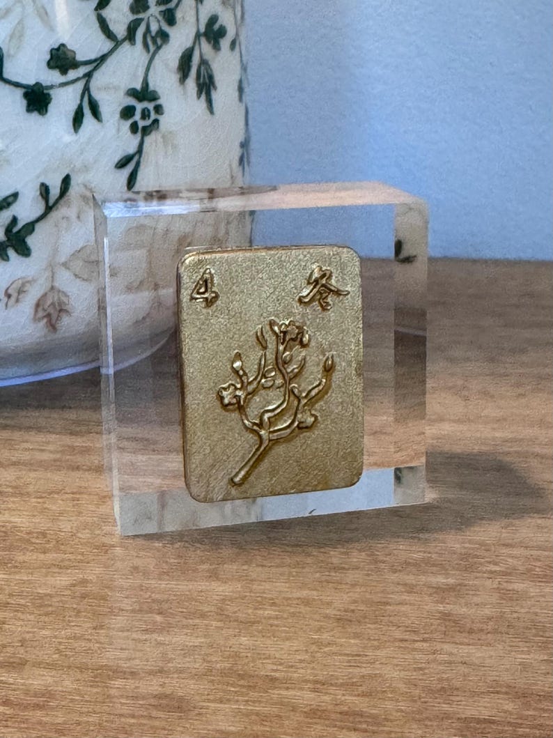 May include: A gold-colored Mahjong tile, featuring the number 4 and Chinese characters, is encased in a clear acrylic block. The tile has a floral design. The block sits on a wooden surface, with a decorative vase in the background.