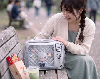 May include: A silver, rectangular display bag shaped like a vintage television set with a clear front window. Inside the bag, a small plush doll with white hair and a dark outfit is visible, accompanied by two iridescent, star-shaped hanging charms.