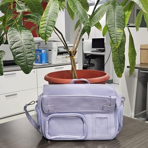 Peut inclure: Un sac messager violet clair doté d'une poignée supérieure, d'un rabat principal et de deux poches avant avec des fenêtres en plastique transparent. Le sac comprend des ferrures en métal argenté, des anneaux latéraux et une fente transparente.