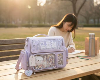 May include: A light purple satchel bag featuring a clear front display window filled with small decorative pins and a white plush cat toy. The bag includes a top handle, an adjustable shoulder strap, and a heart-shaped charm hanging from the side.