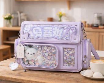 Puede incluir: Una mochila de estilo bandolera de color lavanda con bolsillos transparentes llenos de abalorios y un peluche. La bolsa presenta correas de cadena plateadas, un asa y el texto "喜欢" en blanco. Una etiqueta en forma de corazón cuelga del frente.
