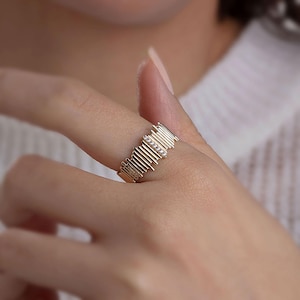 Può includere: Un anello dorato con un design unico a linee verticali e piccole pietre trasparenti. L'anello è indossato su un dito, mettendo in risalto i suoi dettagli intricati e il suo stile elegante. Il design dell'anello è moderno e accattivante.