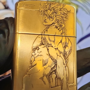 Puede incluir: Un encendedor de metal dorado con una ilustración detallada grabada con láser de un personaje musculoso con cabello puntiagudo. El texto "DIO" está grabado en la esquina superior izquierda y el encendedor tiene una forma rectangular.