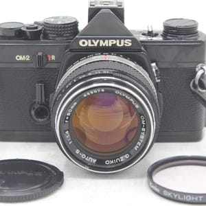 May include: A black Olympus OM-2 film camera with a G.Zuiko Auto-S 50mm 1:1.4 lens attached. The camera features a black leather-textured body, a black lens cap, and a separate Kenko Skylight (1B) 49mm filter. A black camera strap is attached to the sides.