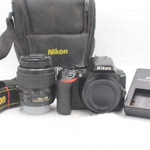 Może przedstawiać: Czarny aparat cyfrowy Nikon D3200 z obiektywem, czarna torba fotograficzna Nikon, pasek do aparatu Nikon z żółtym napisem oraz czarna ładowarka do akumulatorów Nikon MH-24. Korpus aparatu posiada logo Nikon z przodu i na górze.