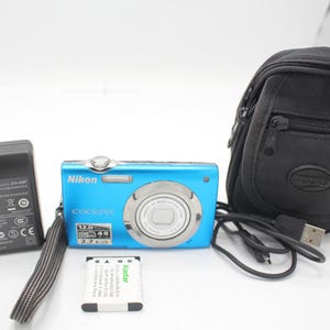 May include: A blue Nikon Coolpix digital camera with a black carrying case, charger, and battery. The camera has a silver lens and the text "COOLPIX" is visible. The camera is accompanied by a black charger and a battery.