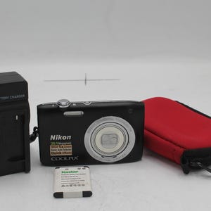 Puede incluir: Una cámara digital Nikon Coolpix negra con lente plateada, funda roja, cargador de batería y batería. La cámara tiene una resolución de 20,1 megapíxeles y zoom 5x. El cargador de batería es negro.