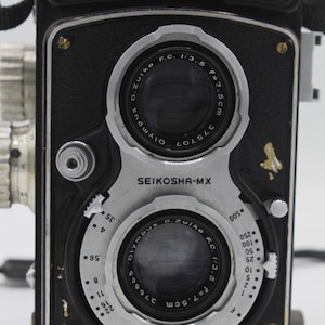 May include: A black Olympus Flex twin-lens reflex camera featuring two D.Zuiko F.C. 1:3.5 f=7.5cm lenses. The front panel displays the text Olympus Flex, Seikosha-MX, and MFX, with silver metal dials for aperture and shutter speed settings.