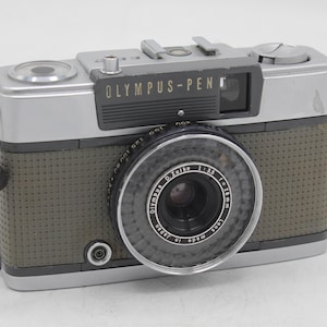 May include: Vintage Olympus Pen camera with a silver and gray body, a textured olive green grip, and a black lens. The camera has the text "OLYMPUS-PEN" on the top and a black strap attached.