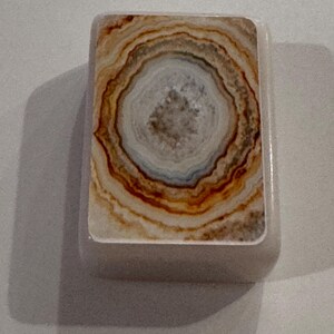 May include: A rectangular, off-white object with a natural stone design. The design features concentric rings of brown, tan, and white, resembling a cross-section of a geode. The object has rounded edges and a smooth surface.