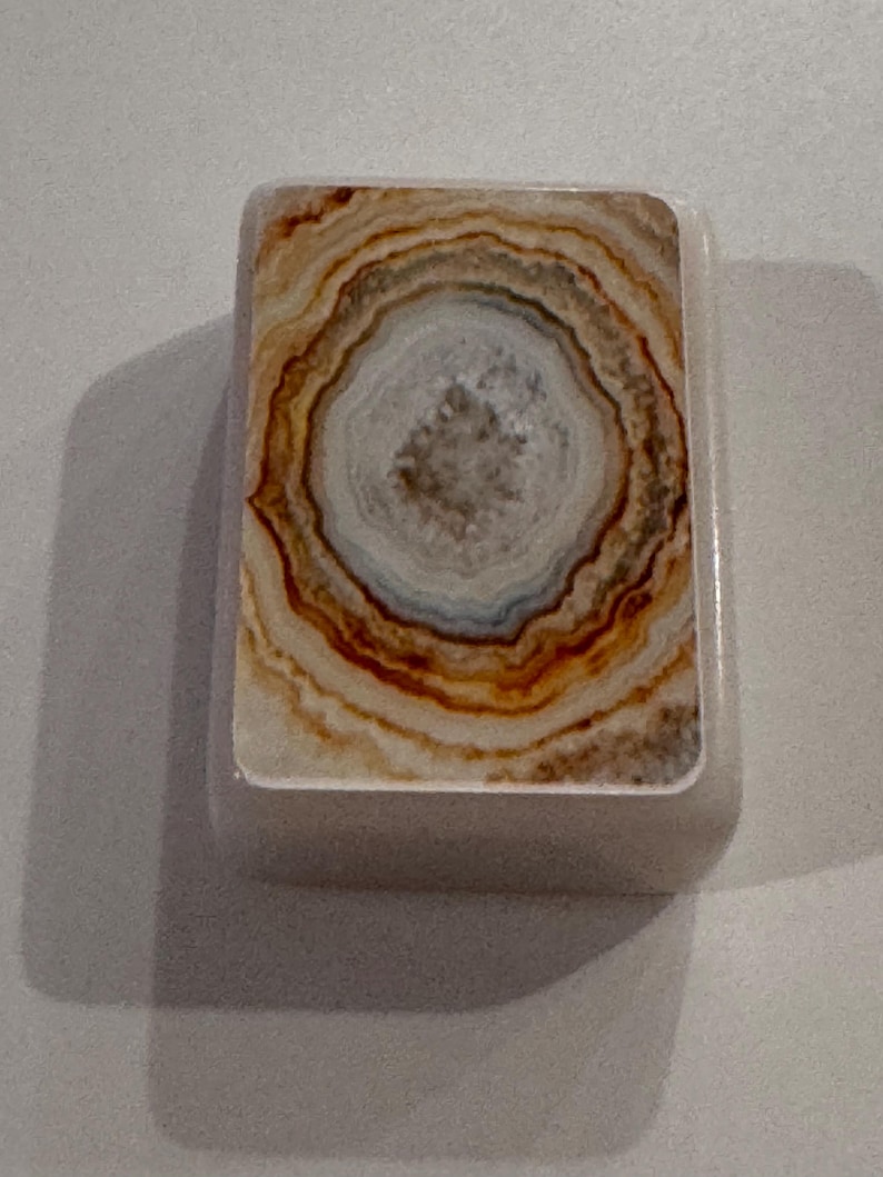 May include: A rectangular, off-white object with a natural stone design. The design features concentric rings of brown, tan, and white, resembling a cross-section of a geode. The object has rounded edges and a smooth surface.