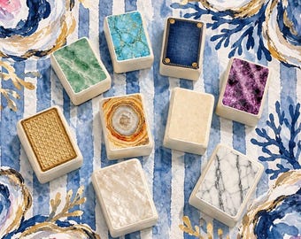 May include: A collection of nine rectangular tiles featuring diverse surface patterns including turquoise stone, denim fabric with gold rivets, green marble, purple amethyst, gold honeycomb, agate, beige stone, white mother-of-pearl, and white marble.