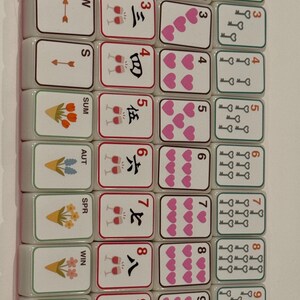 K&ouml;nnte beinhalten: Ein Satz Mahjong-Steine mit verschiedenen Symbolen und Zahlen. Die Steine zeigen Illustrationen von Herzen, Schl&uuml;sseln, Blumen und chinesischen Schriftzeichen. Das Farbschema umfasst Wei&szlig;, Rot und Gr&uuml;n.