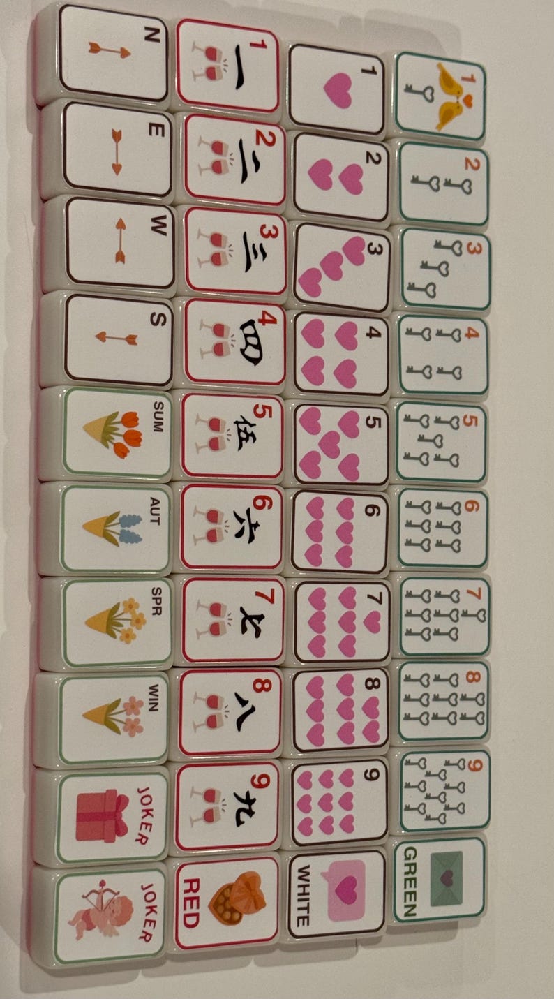 K&ouml;nnte beinhalten: Ein Satz Mahjong-Steine mit verschiedenen Symbolen und Zahlen. Die Steine zeigen Illustrationen von Herzen, Schl&uuml;sseln, Blumen und chinesischen Schriftzeichen. Das Farbschema umfasst Wei&szlig;, Rot und Gr&uuml;n.