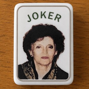 May include: A rectangular white magnet featuring a portrait of a person with dark curly hair, wearing a dark top with a patterned scarf. The word JOKER is printed in dark green capital letters above the portrait, all enclosed in a thin grey border.