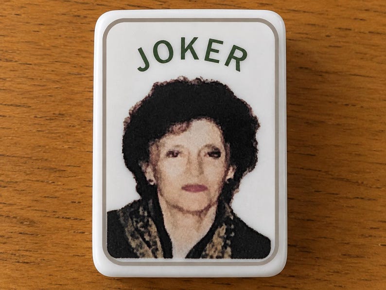 May include: A rectangular white magnet featuring a portrait of a person with dark curly hair, wearing a dark top with a patterned scarf. The word JOKER is printed in dark green capital letters above the portrait, all enclosed in a thin grey border.