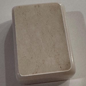 May include: A rectangular, beige soap dish with rounded corners. The dish has a marbled appearance, with subtle brown veining throughout the surface. The edges are smooth and slightly glossy, creating a clean and simple design.