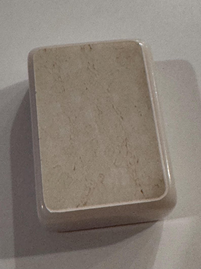 May include: A rectangular, beige soap dish with rounded corners. The dish has a marbled appearance, with subtle brown veining throughout the surface. The edges are smooth and slightly glossy, creating a clean and simple design.
