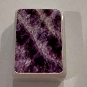 May include: A rectangular object with a marbled purple and white design. The object has a white border and a smooth, glossy finish. The design resembles amethyst or other natural stone.