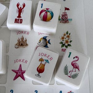May include: Close-up of white game tiles with colorful beach-themed illustrations. The tiles feature images of a lobster, beach ball, cocktail, flamingo, and starfish. Some tiles have the word "JOKER" or a number. The game appears to be a variation of Mahjong.