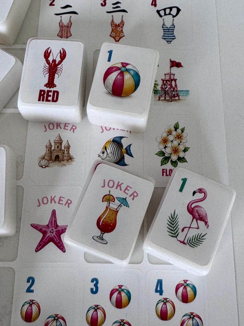 May include: Close-up of white game tiles with colorful beach-themed illustrations. The tiles feature images of a lobster, beach ball, cocktail, flamingo, and starfish. Some tiles have the word "JOKER" or a number. The game appears to be a variation of Mahjong.