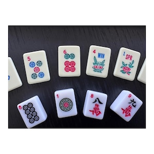 May include: A collection of white Mahjong tiles with various symbols and numbers in red, green, and blue. The tiles are arranged on a dark surface, showcasing the game's components.