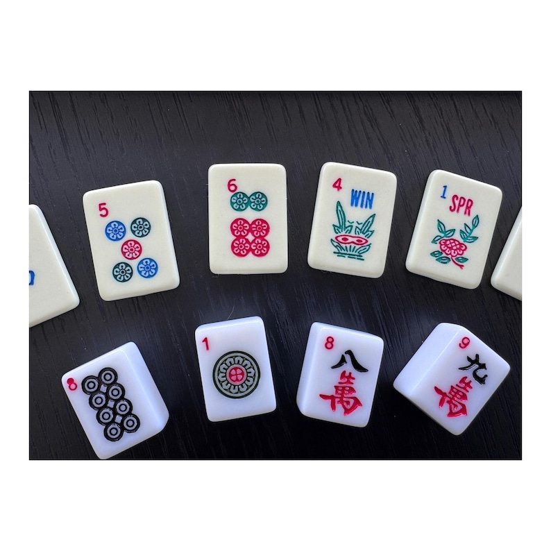 May include: A collection of white Mahjong tiles with various symbols and numbers in red, green, and blue. The tiles are arranged on a dark surface, showcasing the game's components.