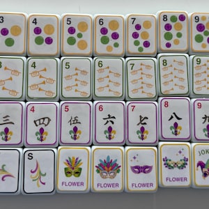May include: A collection of white Mahjong tiles with various symbols, numbers, and characters. The tiles feature purple, green, and gold accents, along with images of a pelican, trumpets, and a crown. Some tiles have the words "WHITE", "GREEN", "RED", and "JOKER".