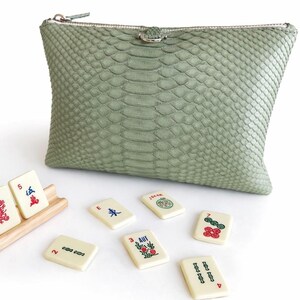 May include: A sage green, textured clutch with a silver zipper and a small silver clasp. Several ivory-colored Mahjong tiles are scattered around the clutch, some in a wooden rack. The tiles have red, green, and blue symbols.