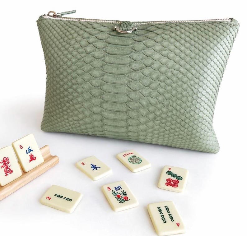 May include: A sage green, textured clutch with a silver zipper and a small silver clasp. Several ivory-colored Mahjong tiles are scattered around the clutch, some in a wooden rack. The tiles have red, green, and blue symbols.